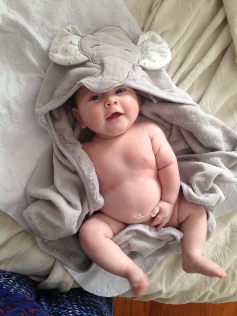 A cute, fat, naked baby wrapped in an elephant bath towel laying on his back on a bed.